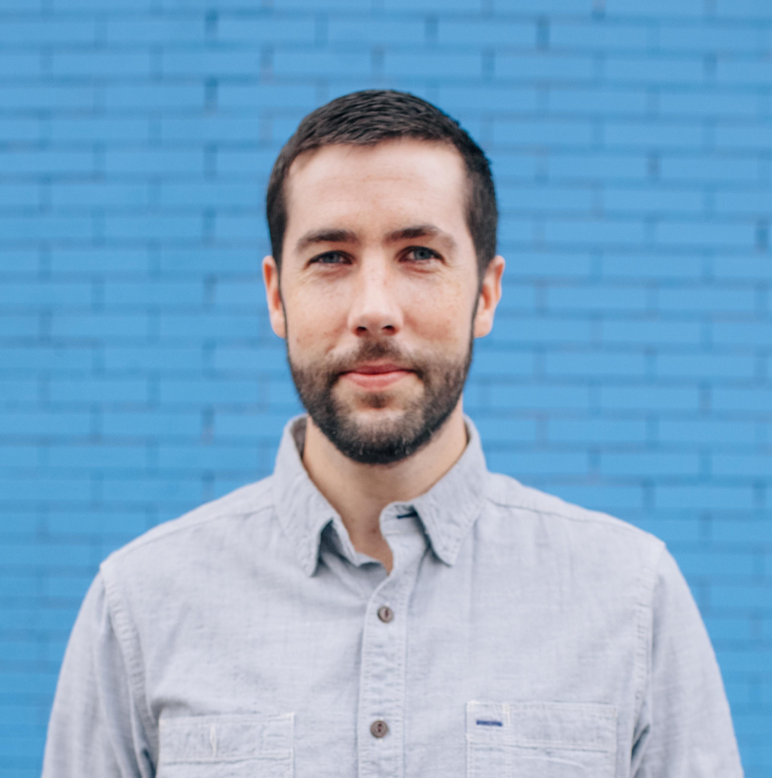 Headshot of David Felix Sutcliffe against a baby blue brick wall.