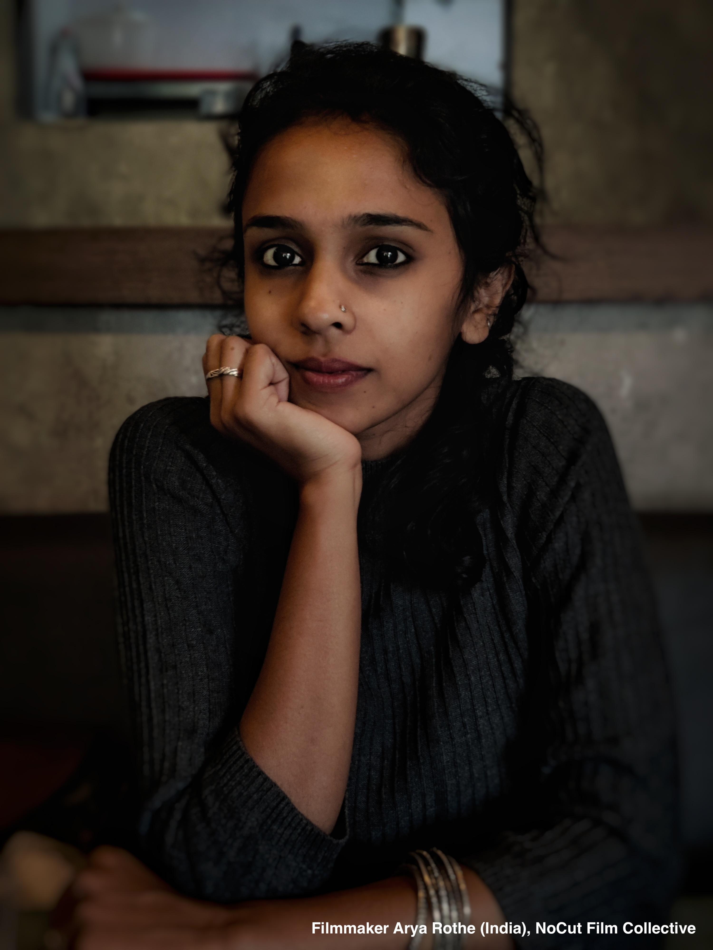 Arya Rothe — Portrait of filmmaker Arya Rothe (India), NoCut Film Collective. A woman with dark hair tied back, wearing a dark grey ribbed sweater, resting her chin on her hand in a dim, warm-toned interior.