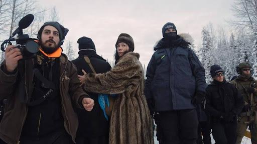 A group of people stand in a line shielding each other from impending violence. A person holds a camera and police in military garb circle.