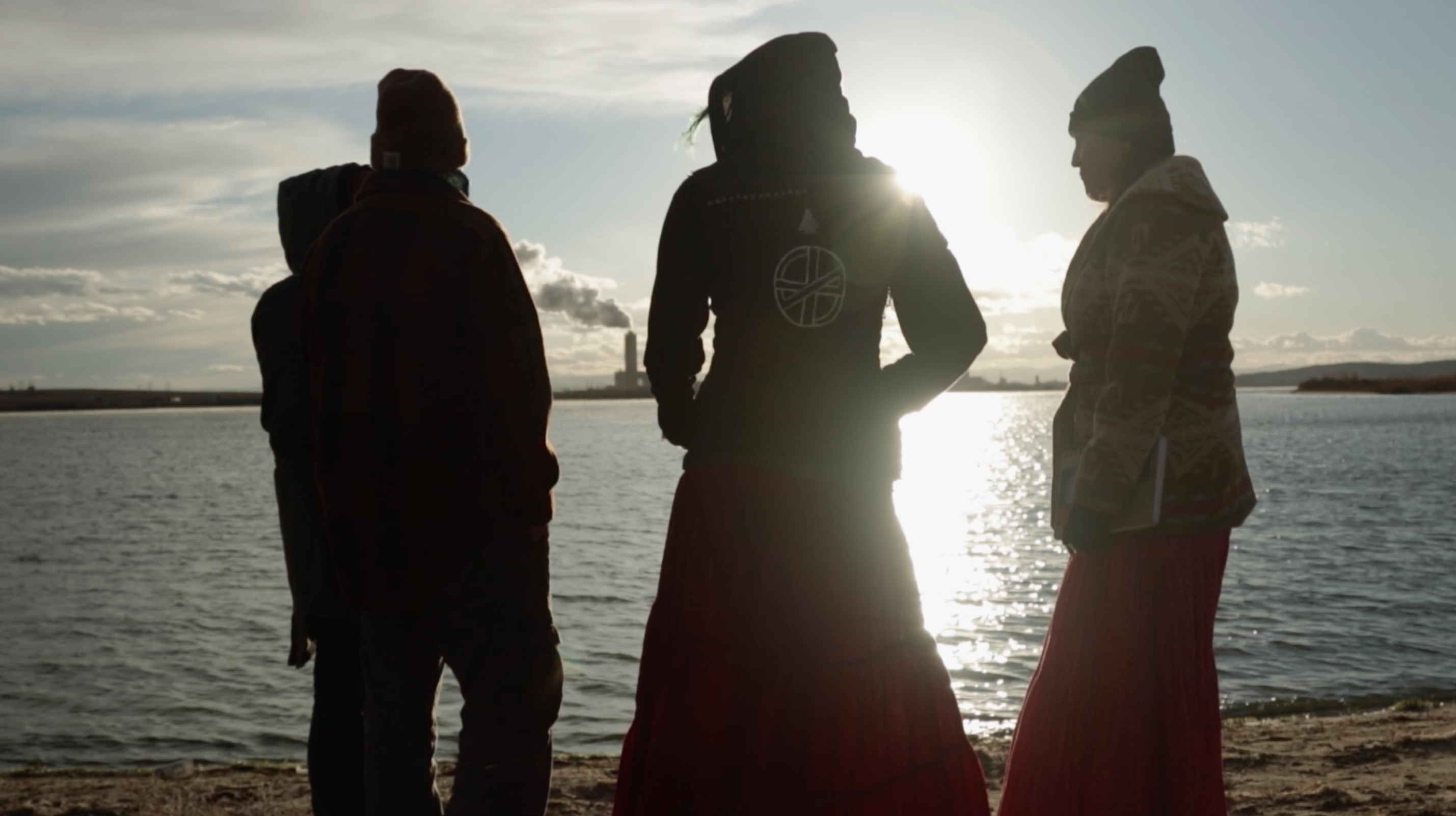 Four people standing on the shore, the sun is setting so we can only see their overshadowed backs, in the background the sea reflects the sun's light