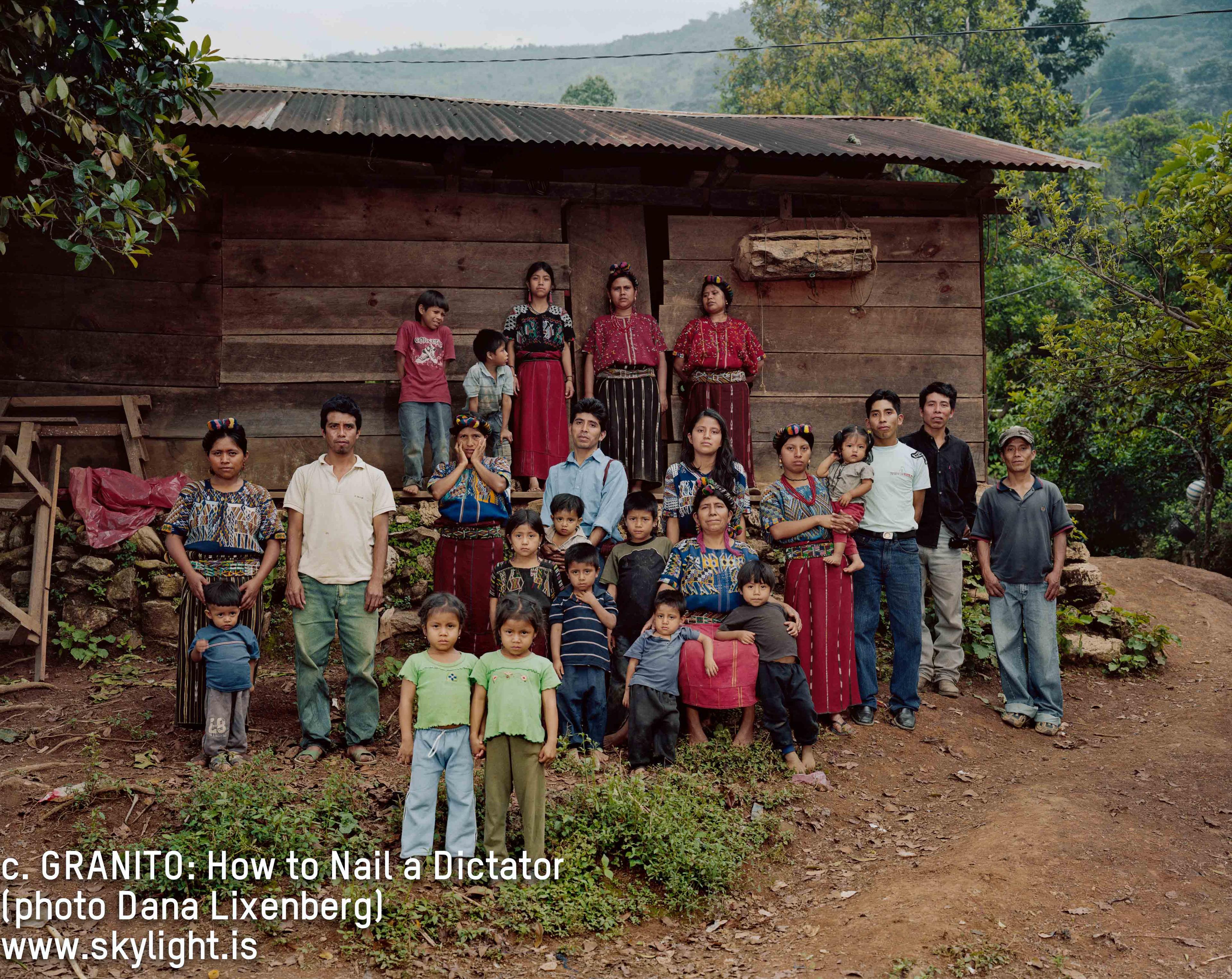 A group of Indigenous Guatemalan people stand in front of a house and look at the camera.