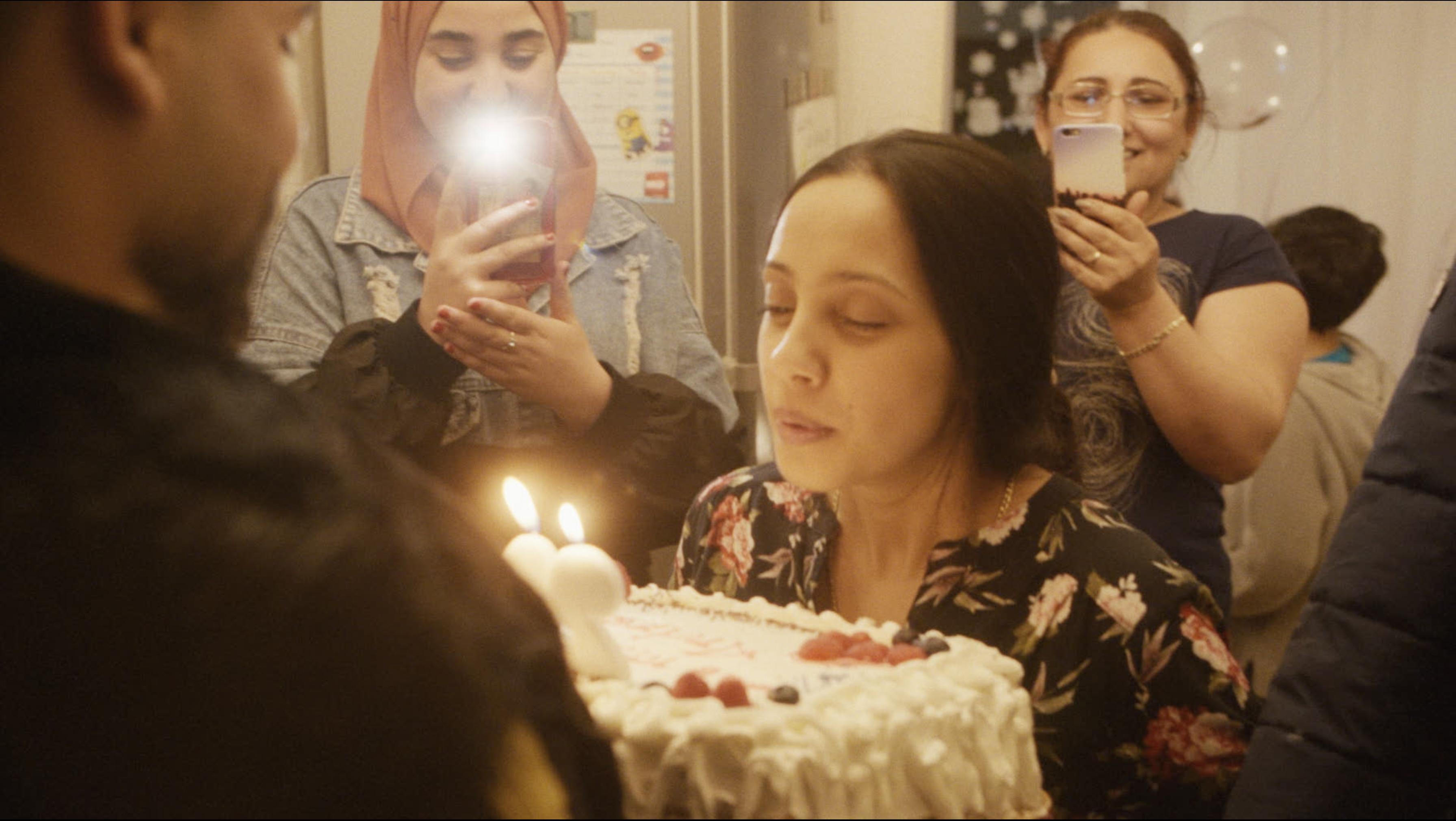 A person blows out candles on a birthday cake while two people behind her record it.