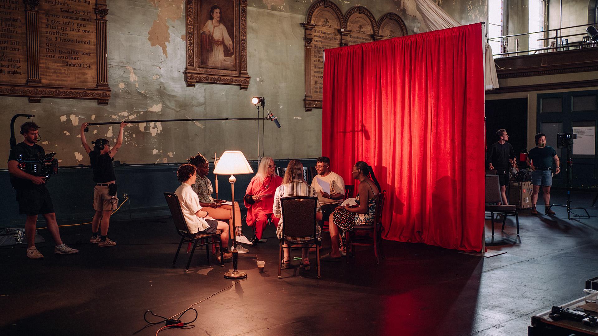 A diverse group of people sit in chairs around a small table, under soft lighting from a nearby lamp, in front of a vibrant red curtain. Film crew members and equipment are visible in the background.