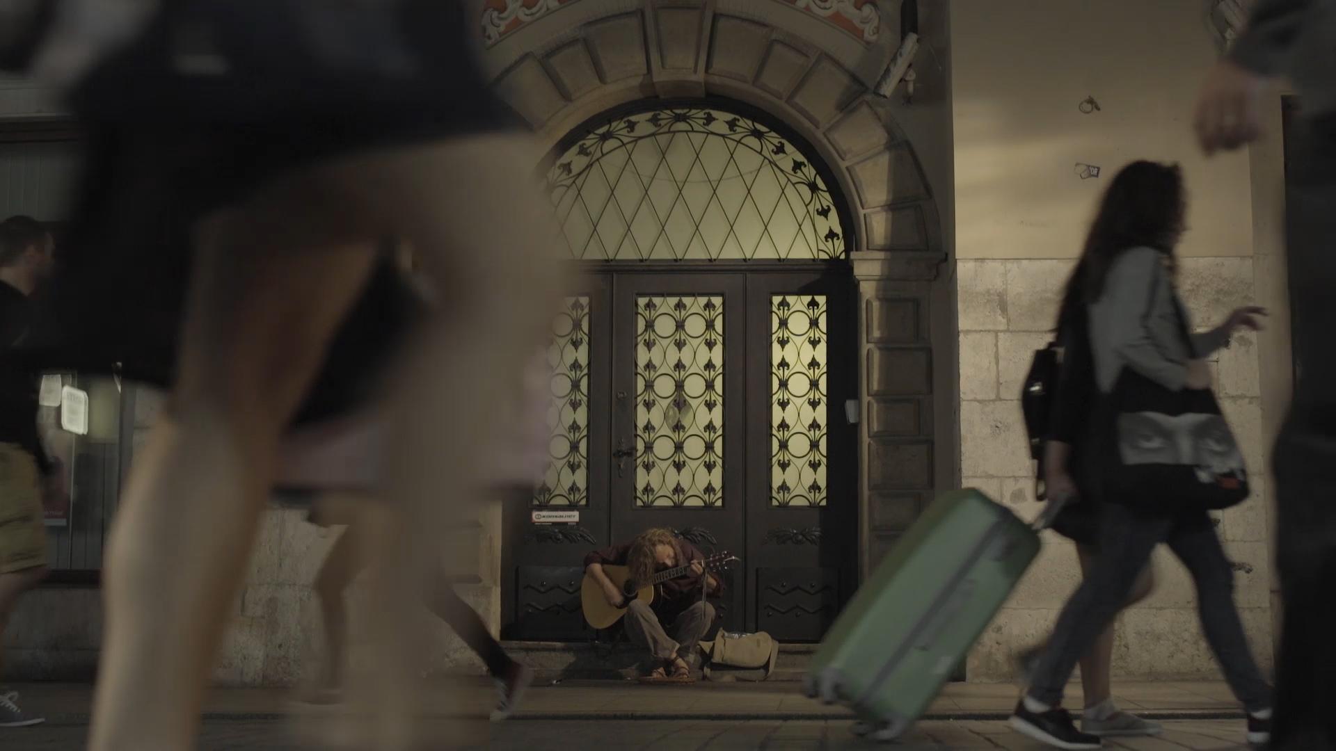 Still from People I Know. A person sitting cross-legged on a building's stoop looking down and playing an acoustic guitar, with three people in motion on the street in front of him