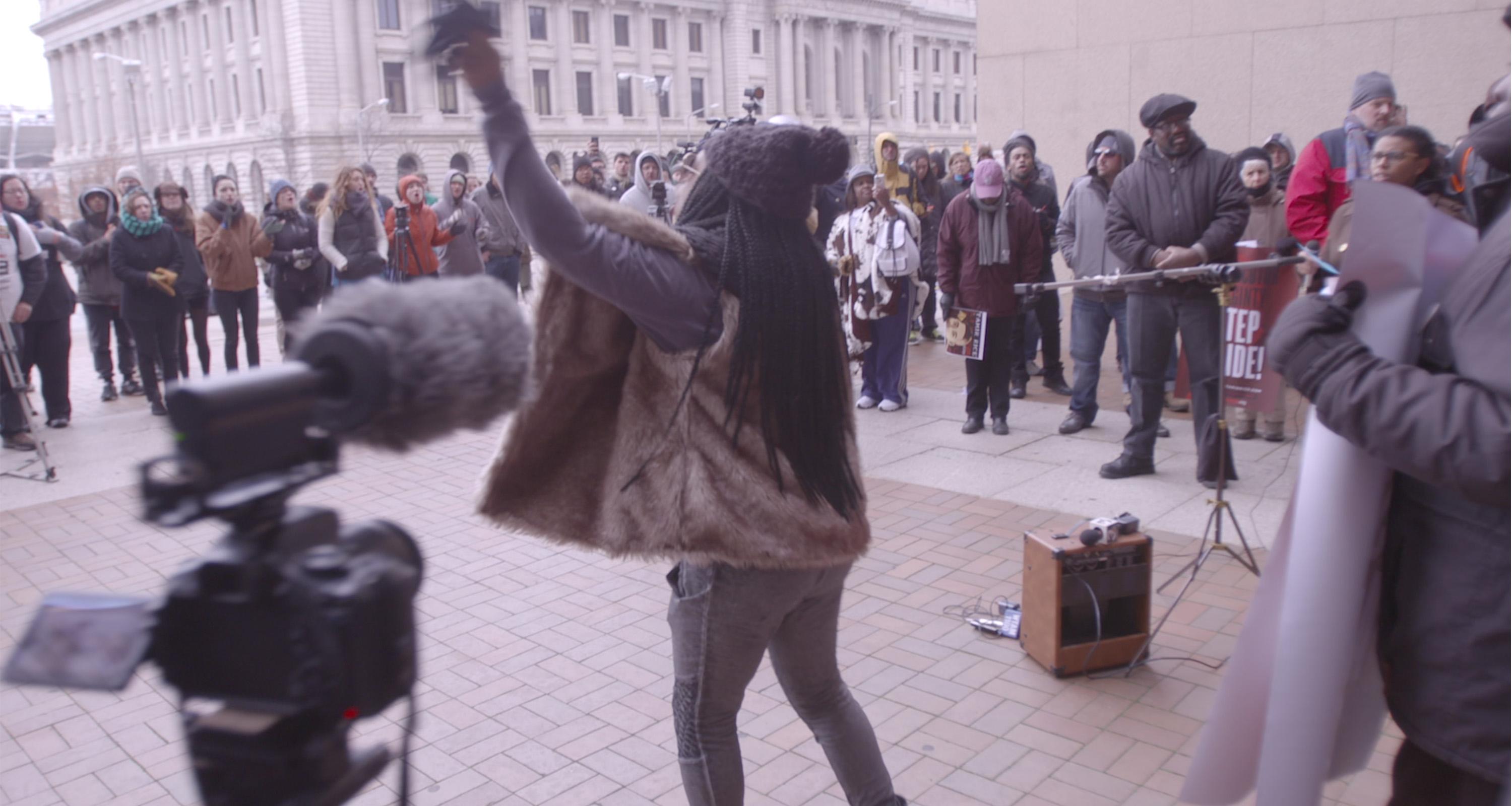 A person leads a protest. They have their hand raised in the air and stand in a circle of other protestors.