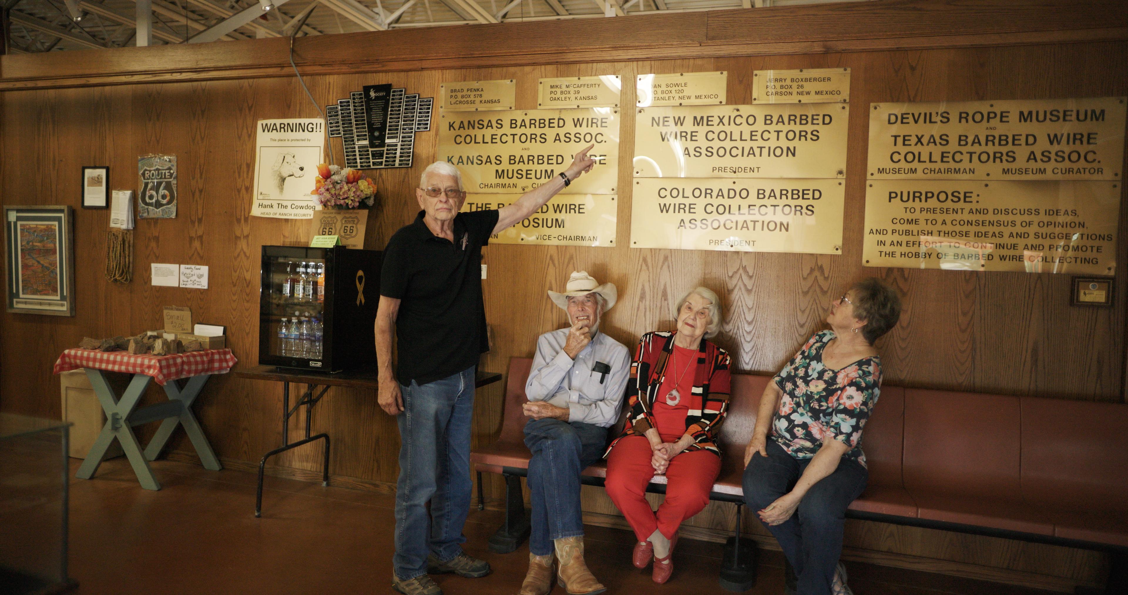 Four older adults sit on a bench inside a barbed wire museum; one man stands pointing to a wall of plaques listing barbed wire collector associations.