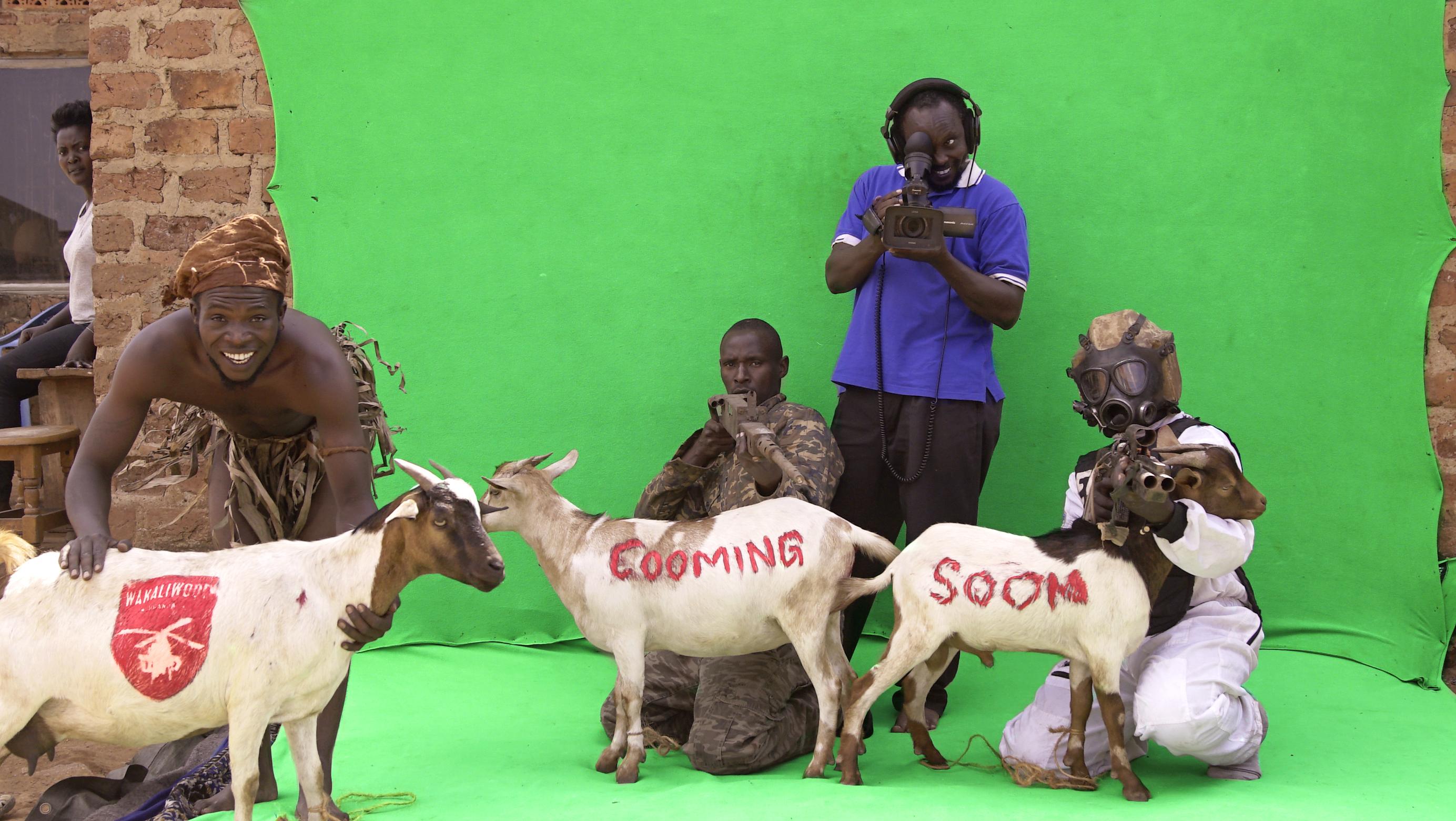 Still from Once Upon a Time in Uganda. Four men in front of a greenscreen outside. One is standing, holding a camera. Two men kneel on the ground holding guns and two goats, the words "Cooming soon" are painted in red on the two goats. Another man crouches above a goat with a red logo painted on it.
