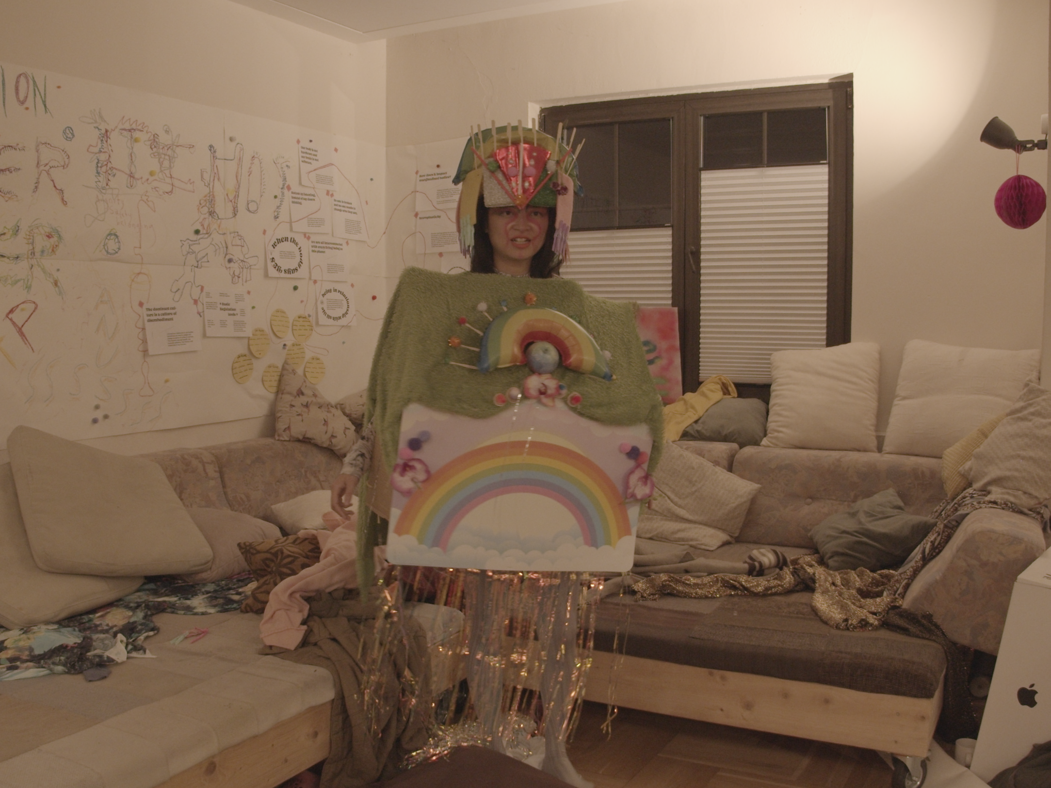 In a messy lounge room, Viv stands alone, looking confused, in a rainbow house costume made out of cardboard.