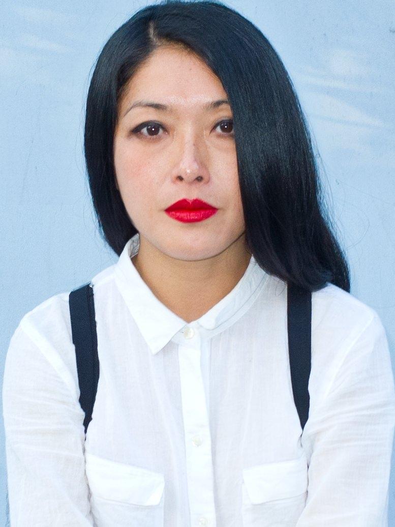Neary Adeline Hay looking directly at the camera. She has long dark hair, and is wearing a white collared shirt, with black suspenders.