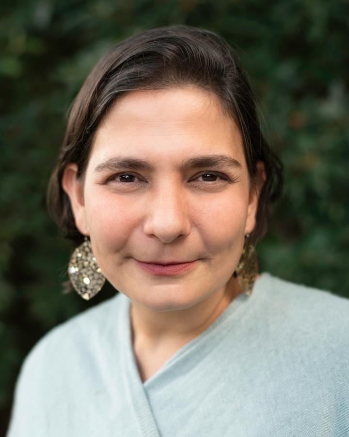Eliza Licht looking directly at camera. She is wearing earrings and is in front of an out focus background.