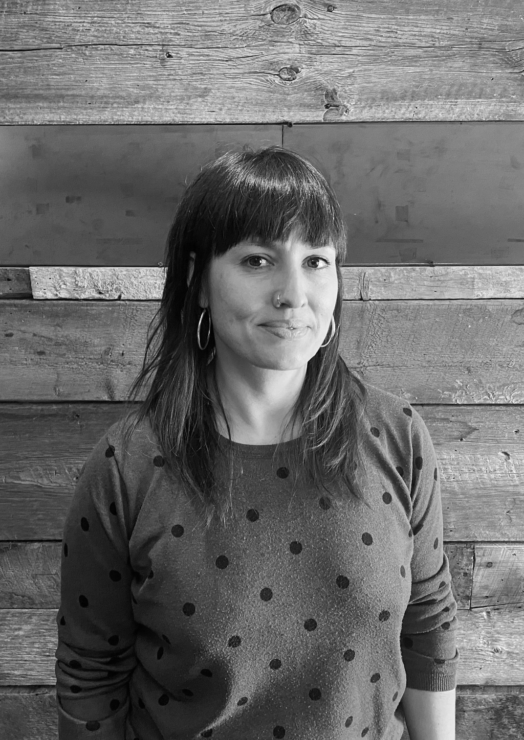 Rocío Barba Fuentes looking directly at camera. She is standing in front of a wooden paneled wall. She has hazelnut eyes, dark, medium length brown hair, a nose ring, silver earrings and is wearing a blue sweater with black dots. Black and white portrait.