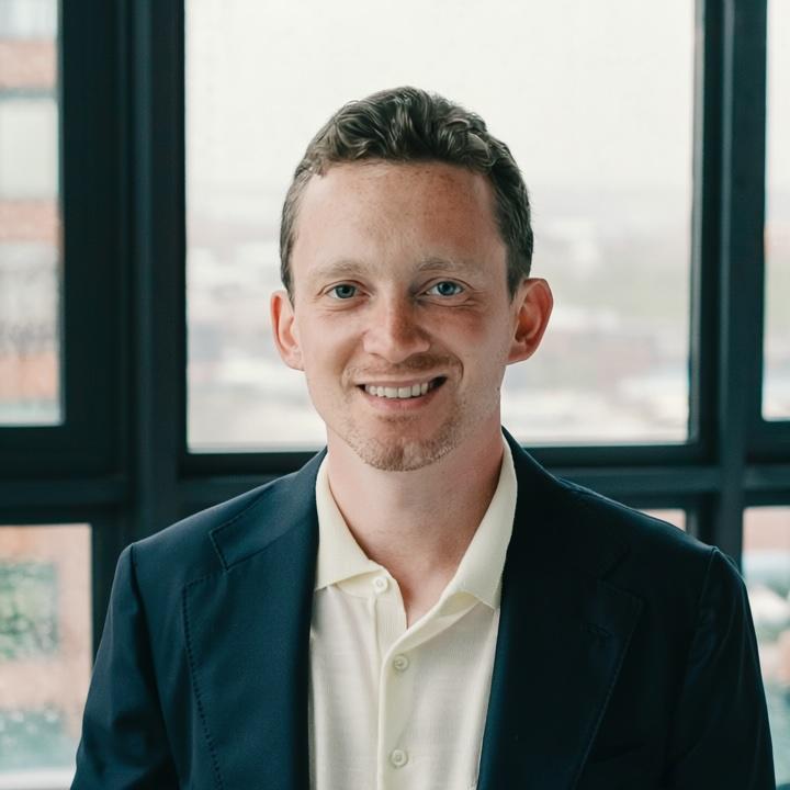 A smiling individual in a dark suit and light shirt stands in front of large windows with a blurry cityscape view.