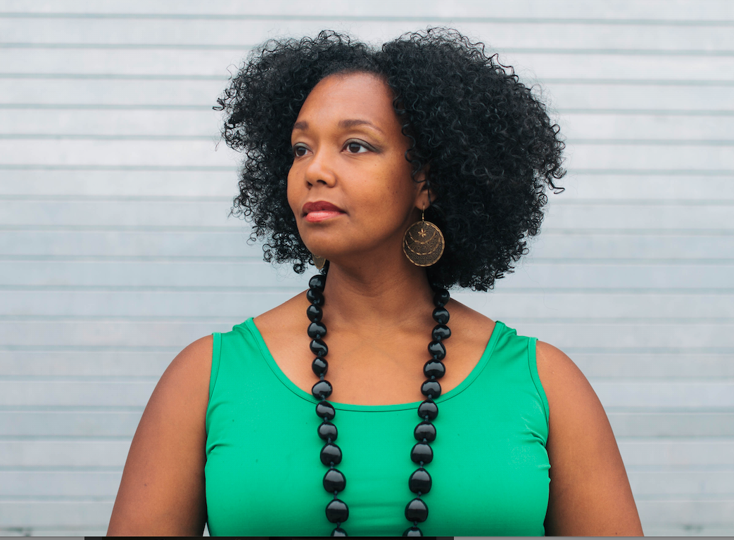 Portrait of Kiara C. Jones, a woman looking to the right, away from the camera, with curly medium hair, wearing a no-sleeve tank, a long necklace and round big earrings.