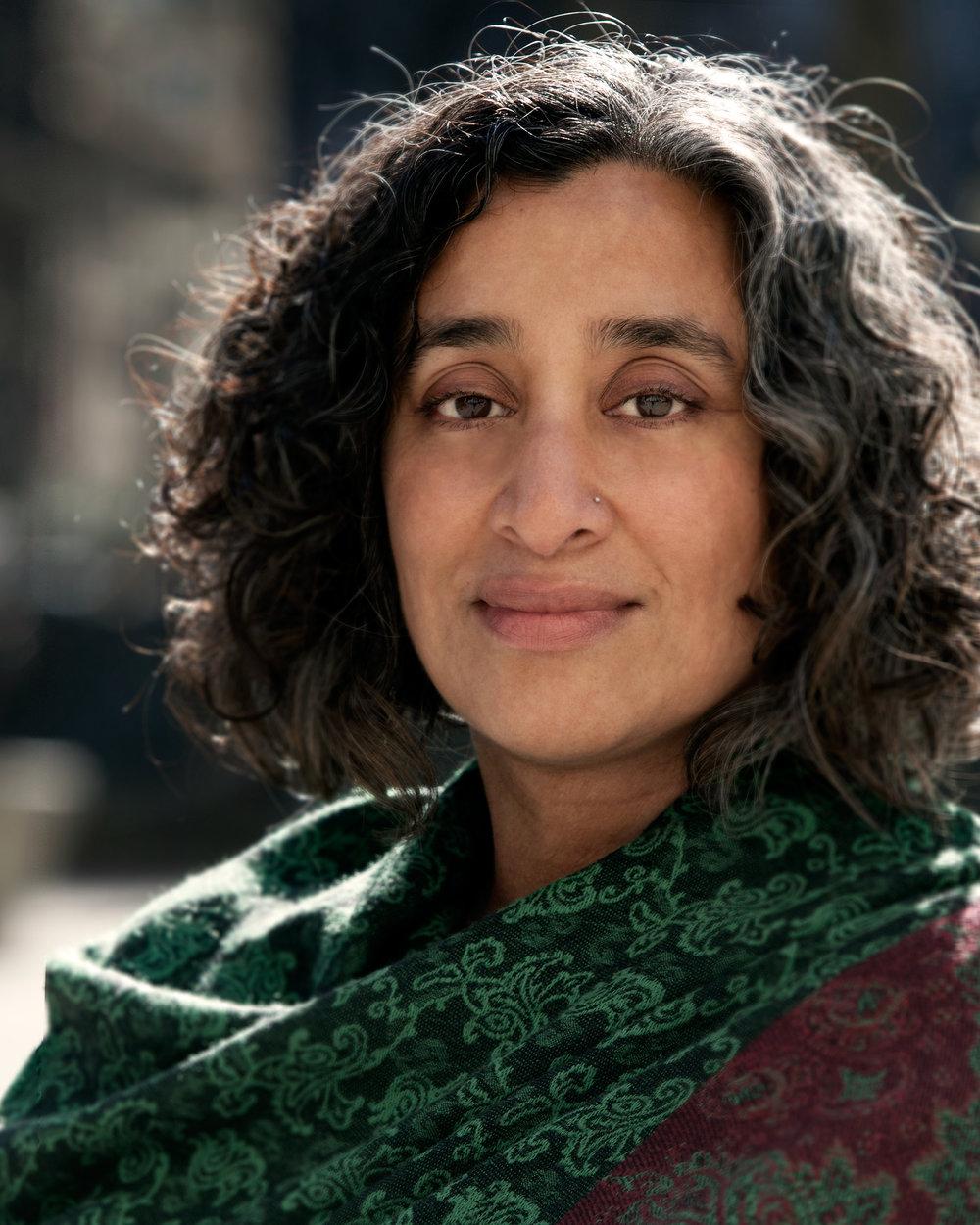 Geeta Gandbhir has medium-length dark wavy hair, wears a scarf, and smiles at the camera.