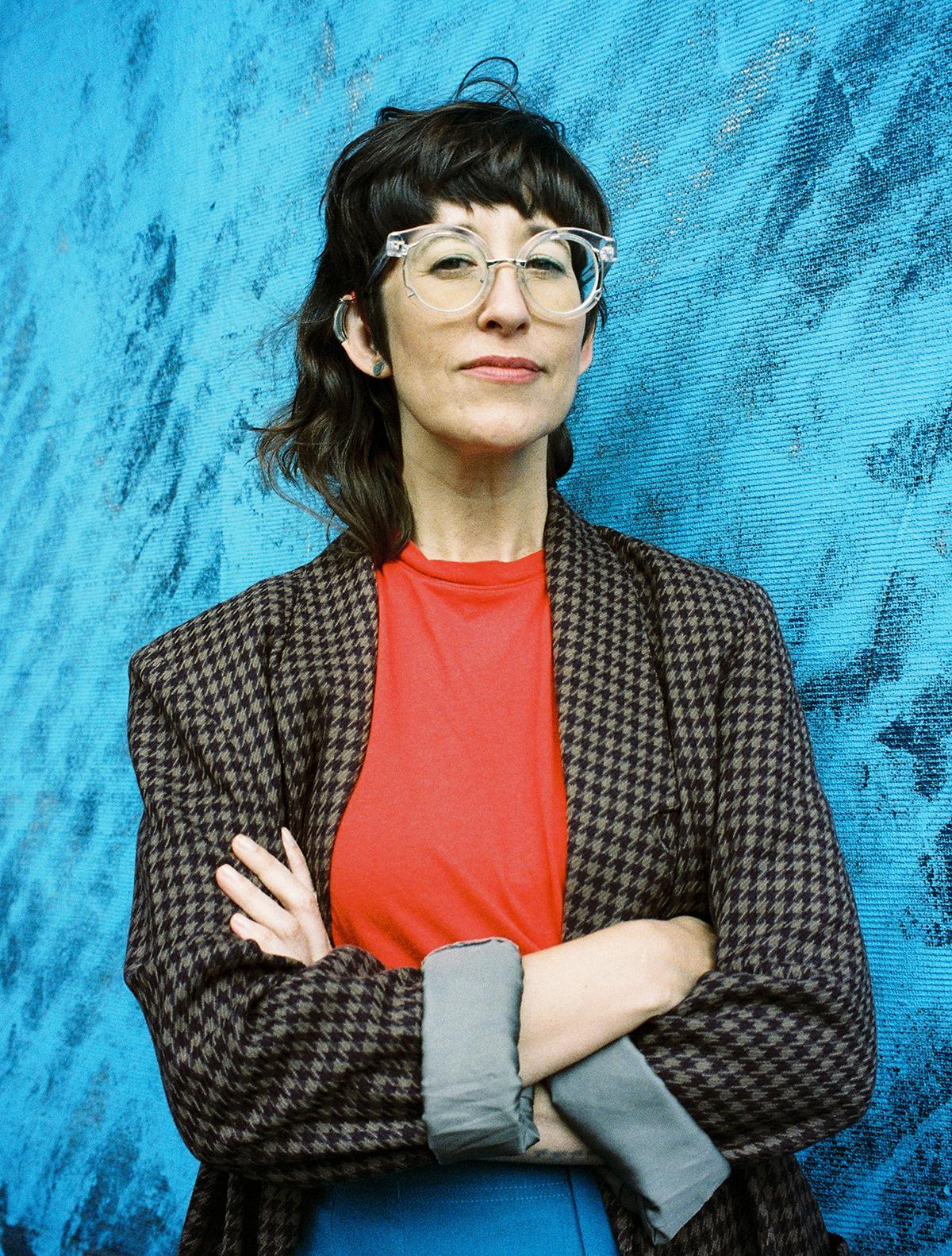Alison O'Daniel is looking directly at the camera. She wears a checkered blazer and red shirt against a blue wall.