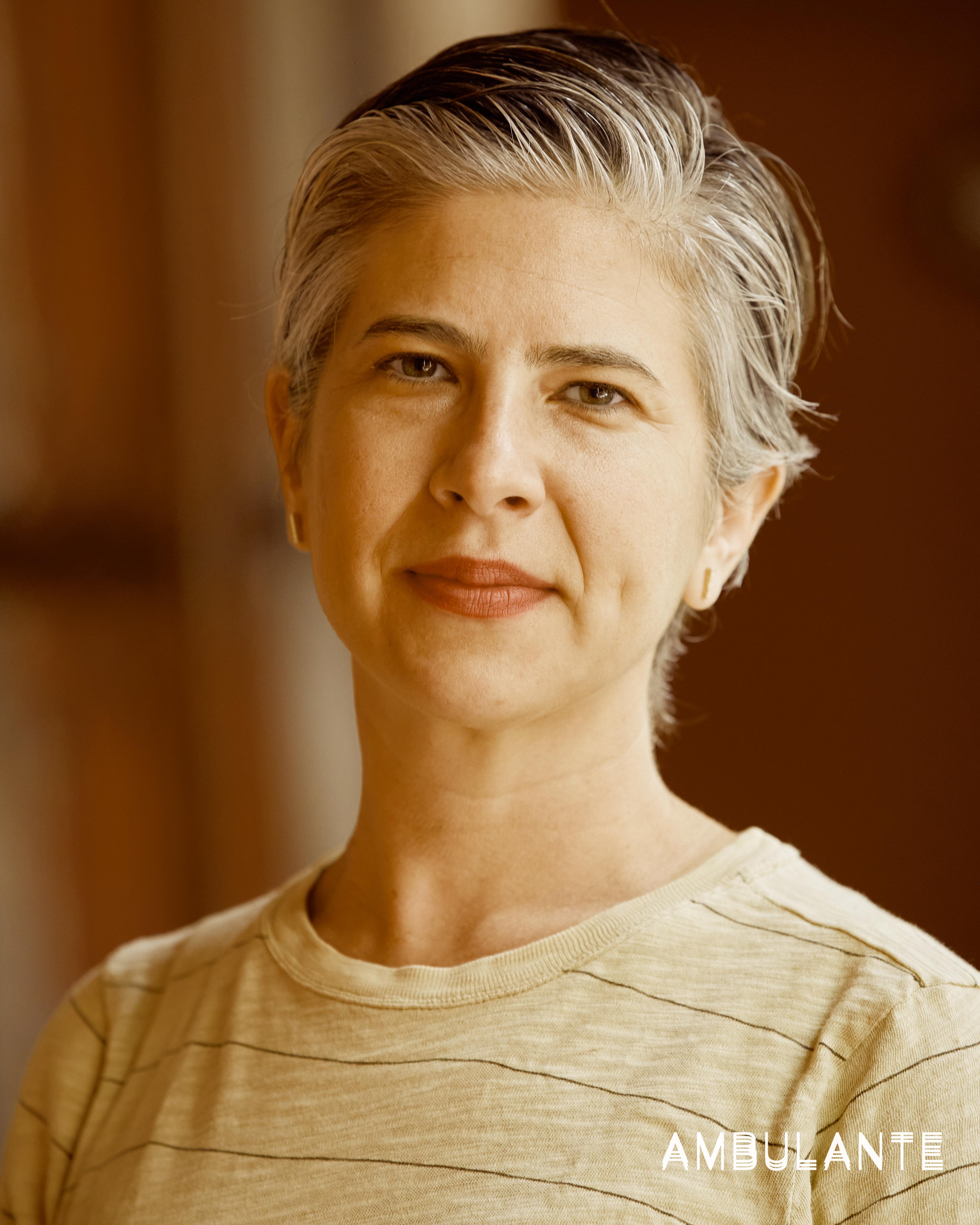 Emily Cohen Ibañez looking directly at the camera. She has short grey and dark brown hair and is wearing gold earrings, lipstick, and a striped T-shirt.