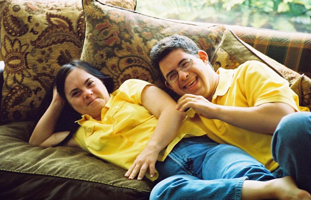 A photo of a couple with Down Syndrome wearing matching yellow shirts laughing on a couch together.