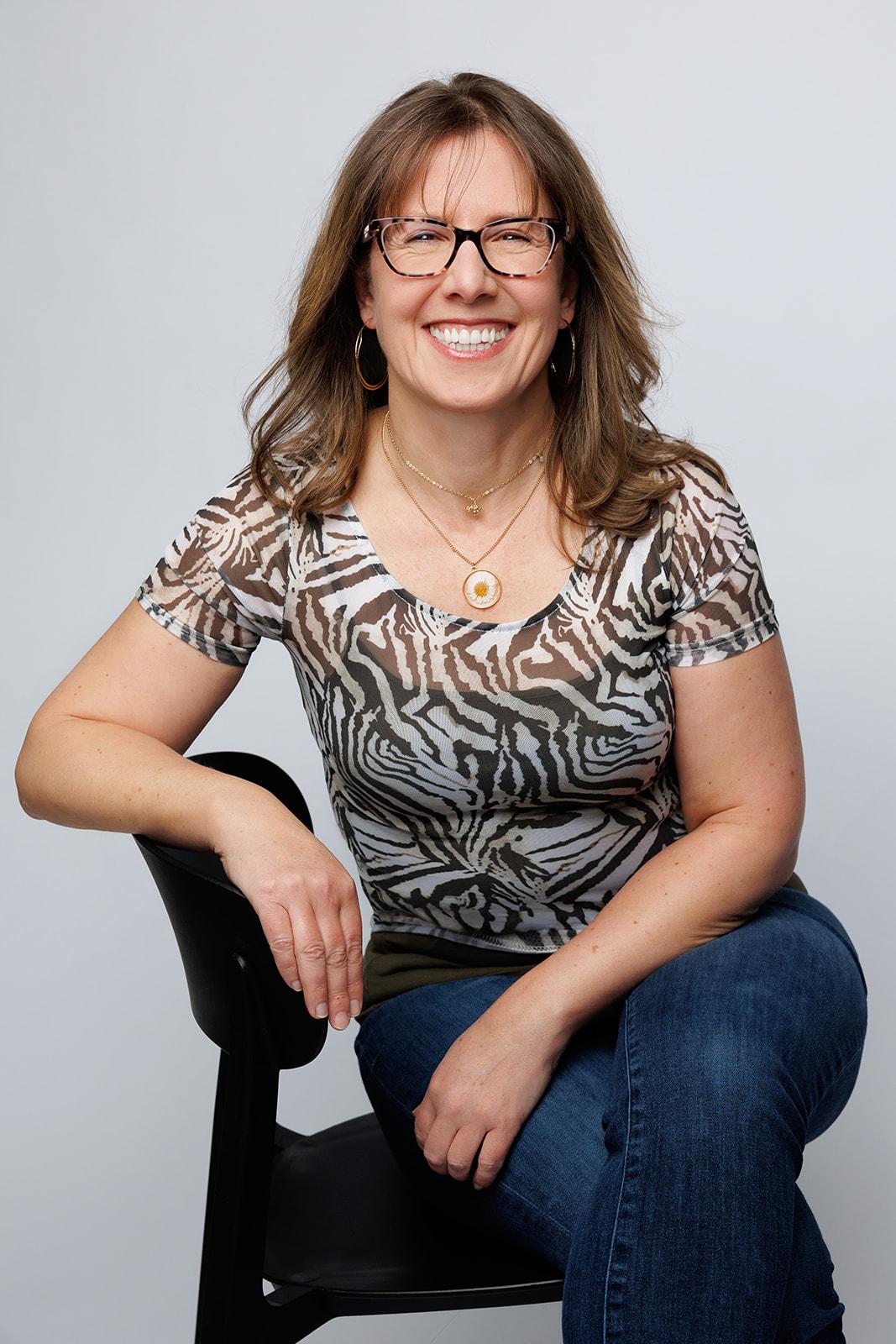 White woman with shoulder-length brown hair and brown glasses leaning on a chair. Headshot of Margo Guernsey.
