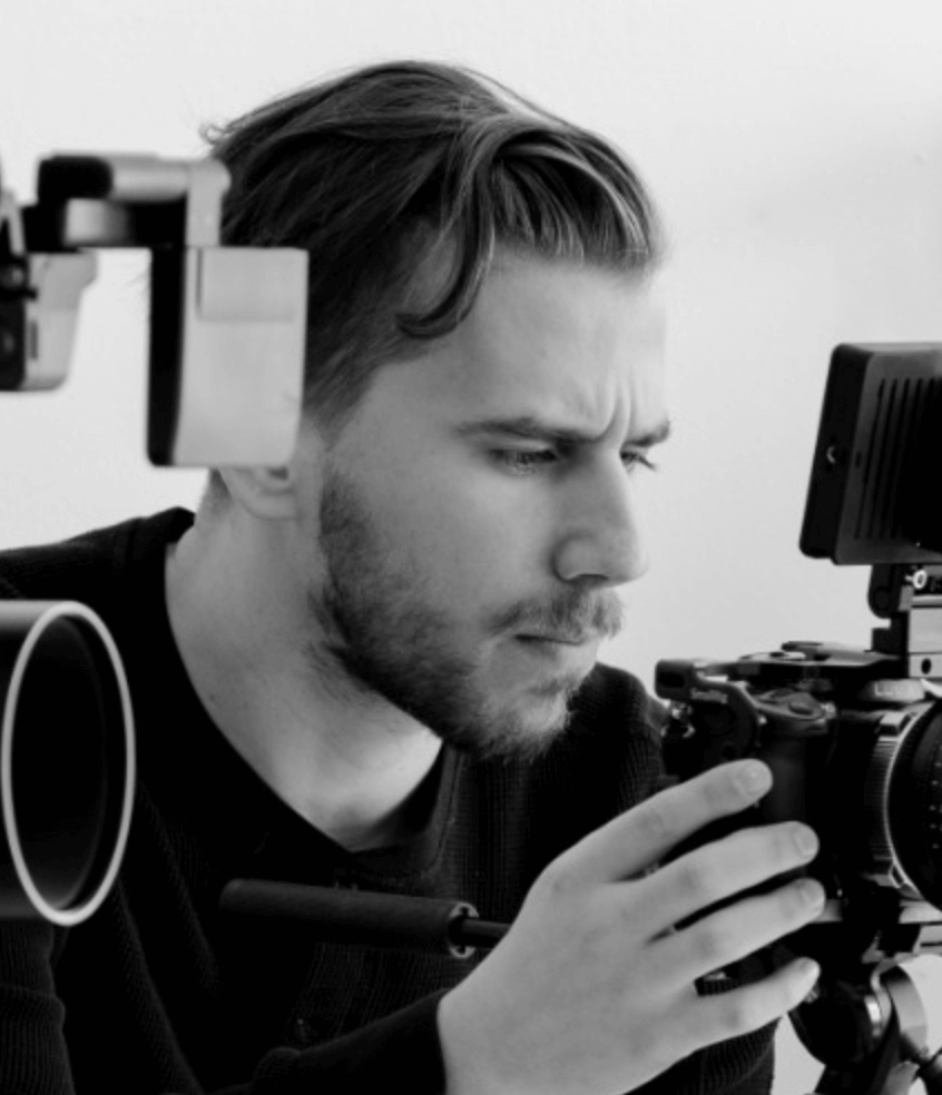 A black and white portrait of Daniel Nelson. A white man with a beard and mustache is surrounded by film cameras, he looks down to the right through a camera lens he holds.