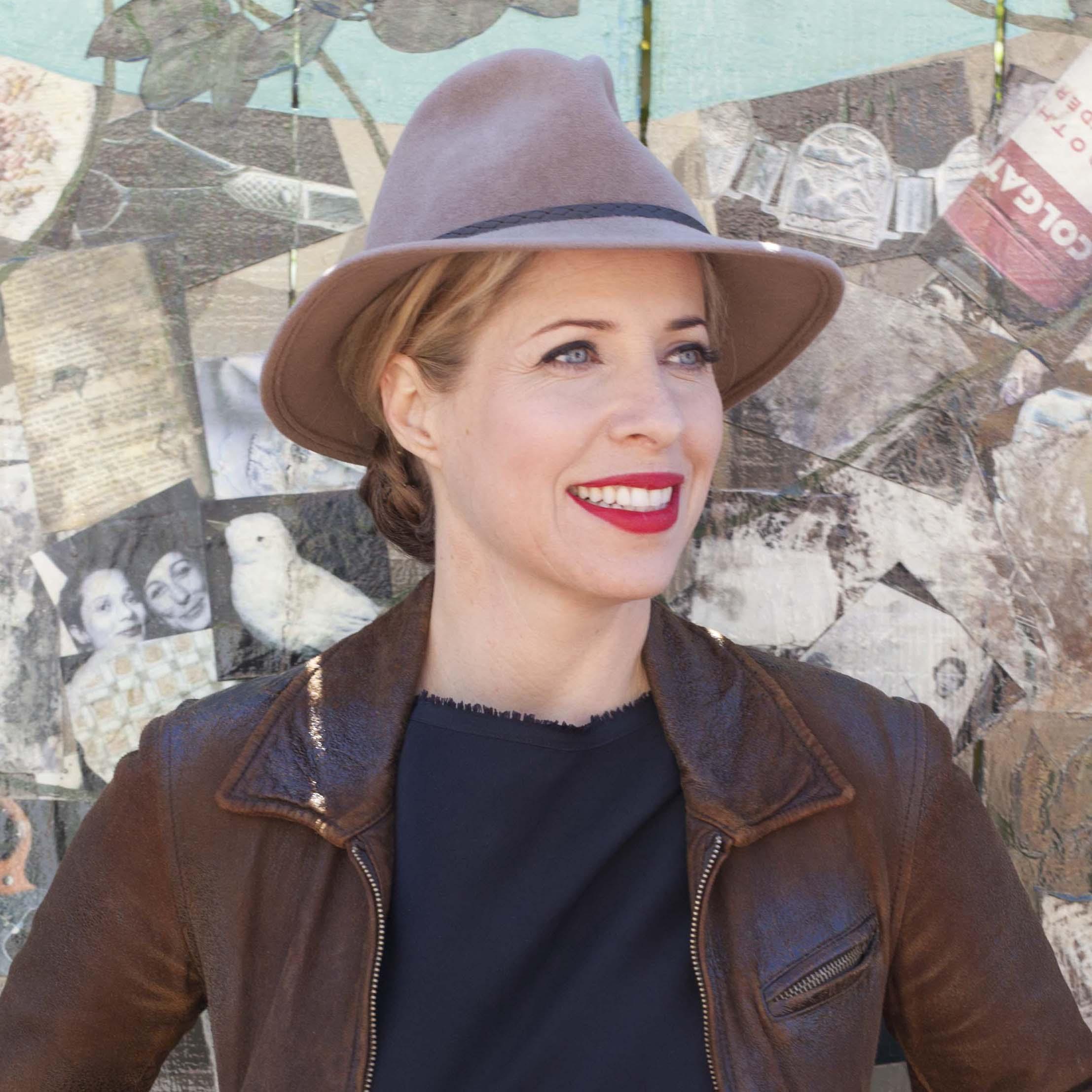 A portrait of a woman wearing a fedora and a leather jacket standing in front of a collage wall. She smiles and looks off camera.