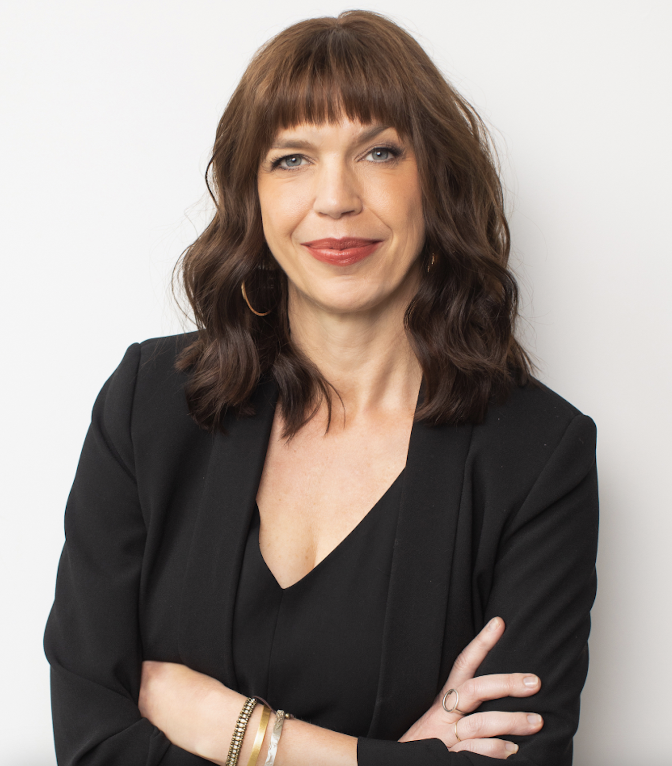 White woman with medium-length brown hair with bangs wearing a black top and blazer, looking directly into the camera while crossing her arms