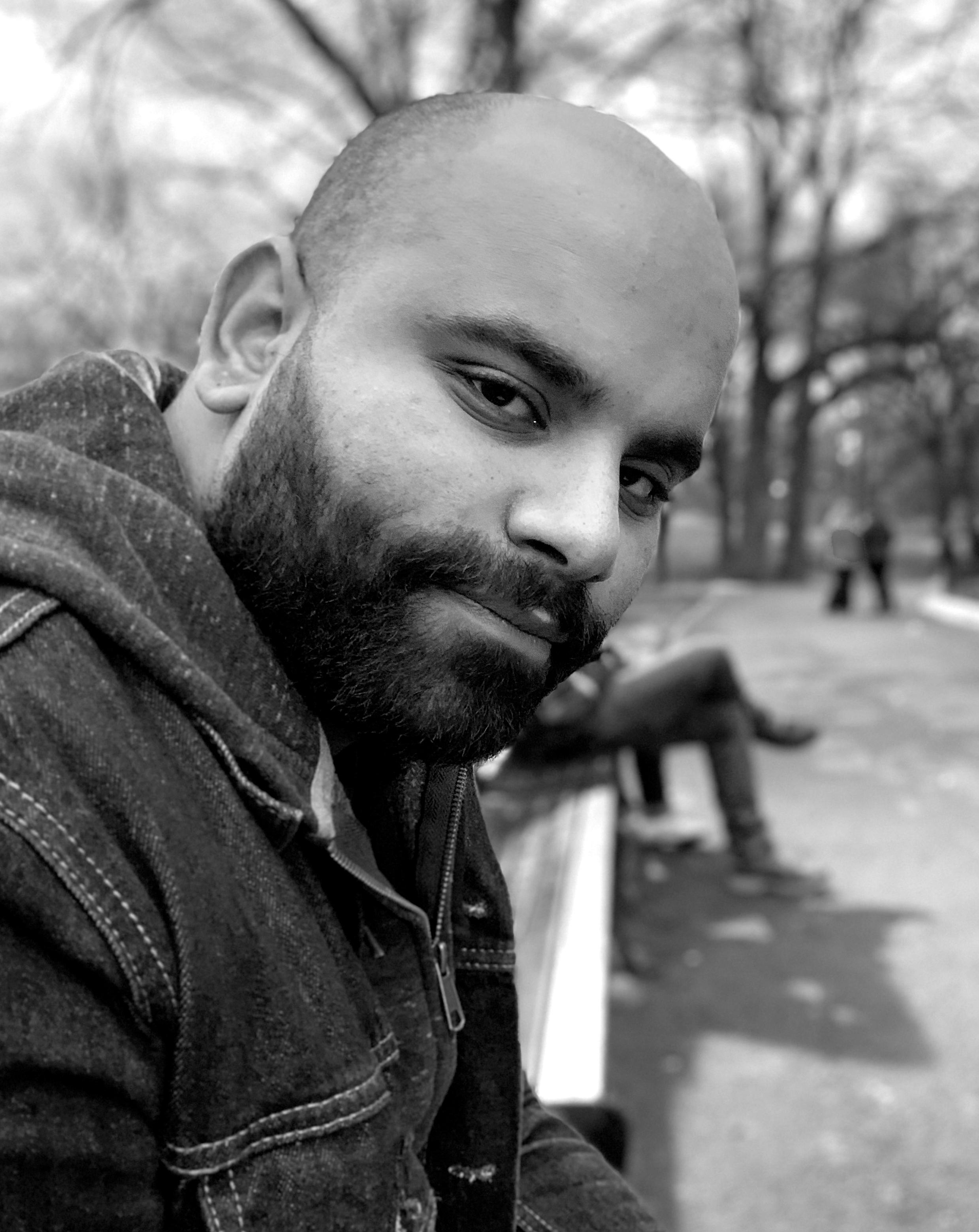 Ronald Baez looking directly at the camera. He is sitting on a bench and is leaning forward. He has a full beard and is wearing a hooded jacket. A figure can be seen seated in the out of focus background. Black and white portrait.